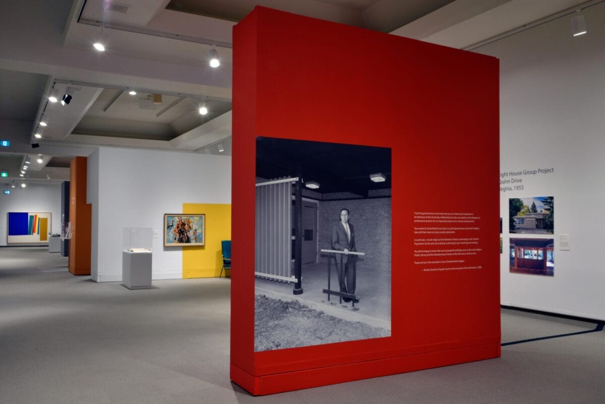 La salle d'exposition d'un musée est dotée d'un grand mur rouge avec une photo en noir et blanc d'une personne debout à l'extérieur d'un bâtiment moderne. La salle comprend diverses œuvres d'art encadrées sur des murs colorés et des vitrines. Des plafonniers illuminent l'espace.