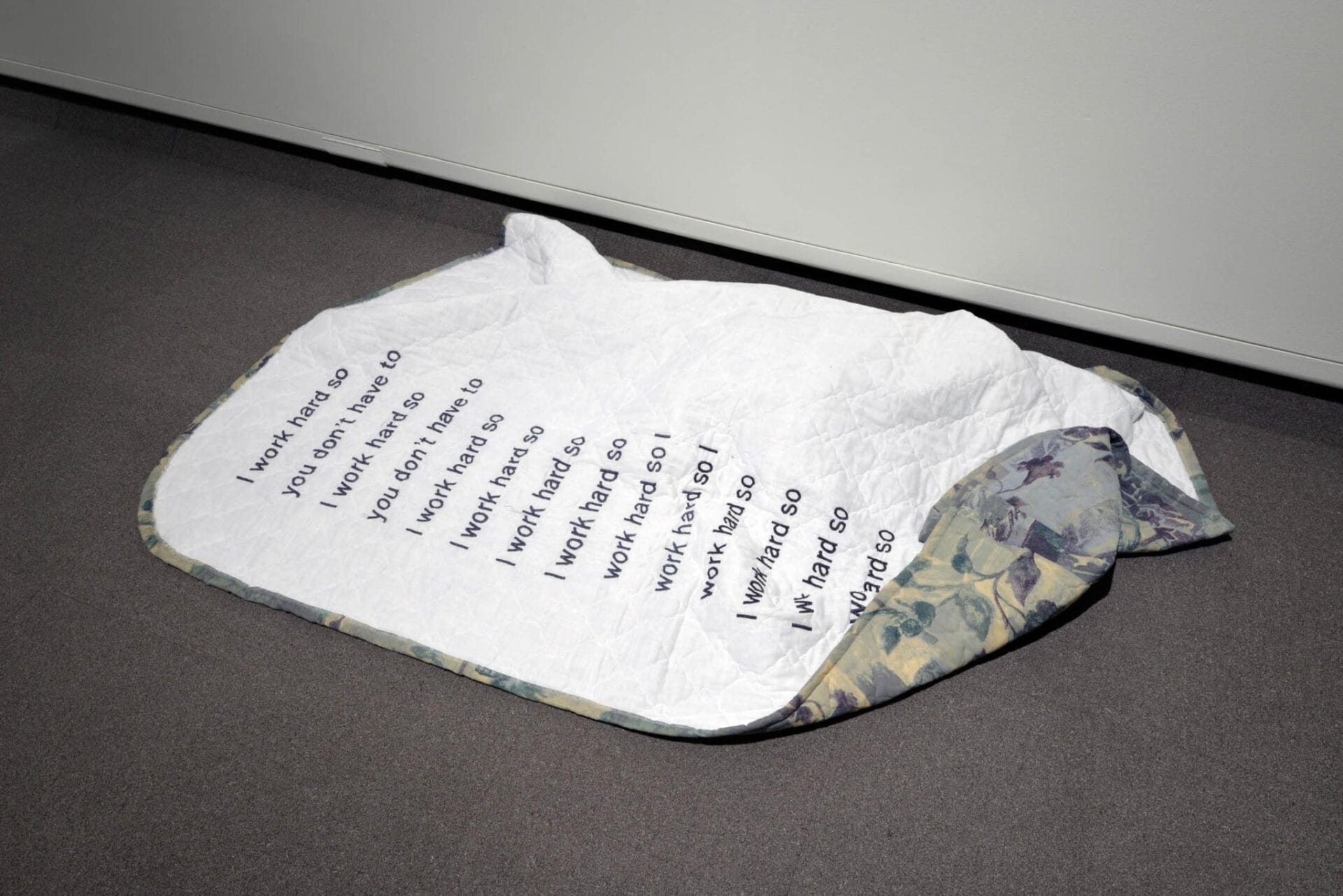 Une couette blanche repose sur une moquette grise contre un mur blanc. La courtepointe comporte un texte répété en lettres noires indiquant : « Je travaille dur donc je travaille dur donc je travaille dur donc » dans un motif vertical. Les bords de la courtepointe ont un motif floral.