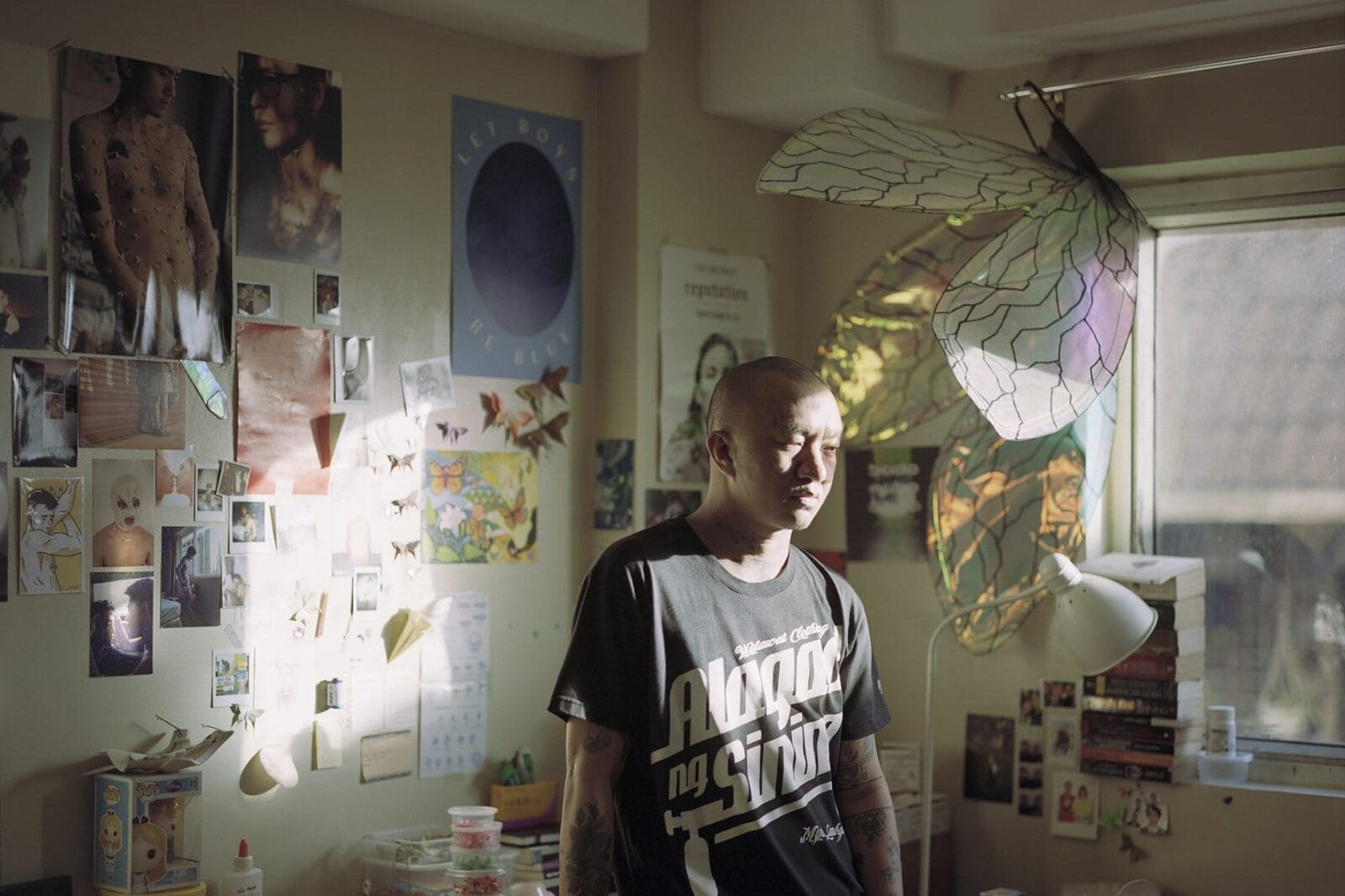 Person with tattoos standing in front of a wall with various posters and clippings of artwork hung up.