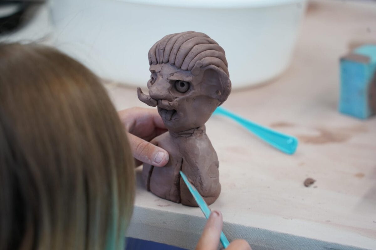 Une personne sculpte une figurine en argile aux traits exagérés, notamment une large moustache et des cheveux texturés, à l'aide d'un outil de sculpture bleu. La figurine est posée sur une table.