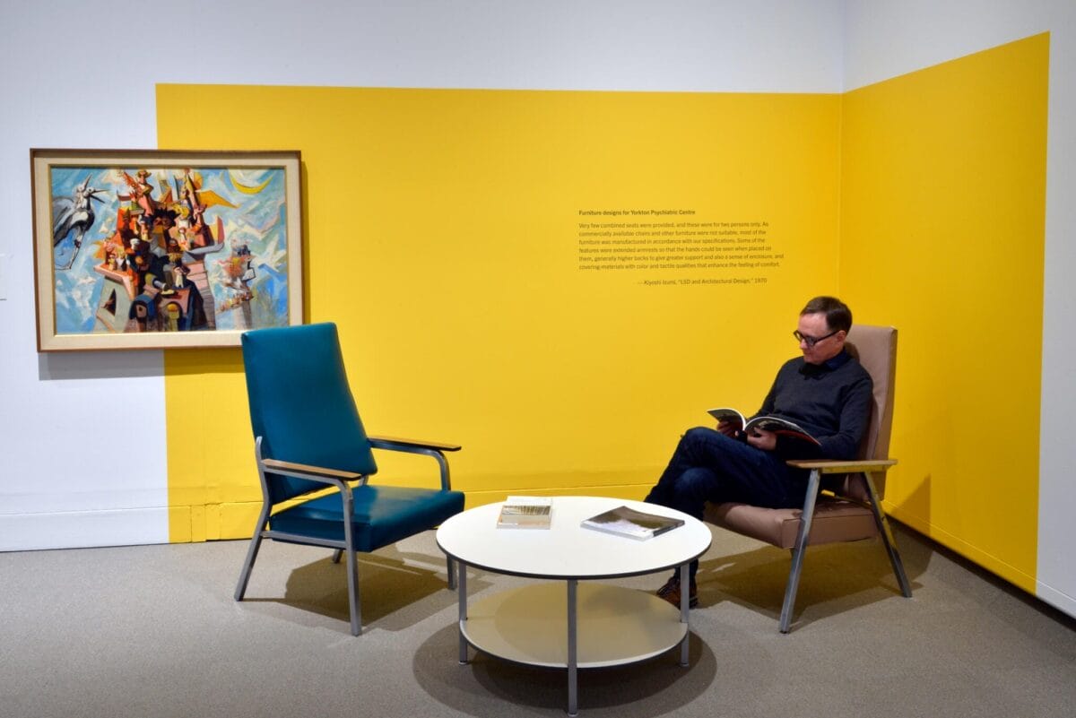 Une personne est assise sur une chaise, en train de lire, dans une galerie aux murs jaune vif. Un tableau d'art moderne est exposé sur le mur. Une autre chaise et une table ronde avec des livres se trouvent devant. L'atmosphère est calme et contemplative.