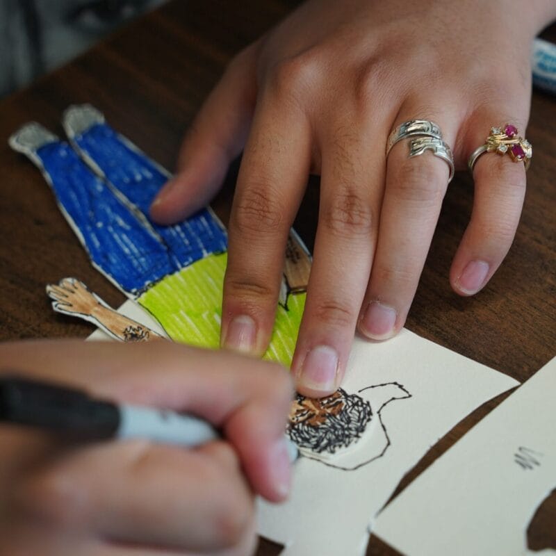 A person colors Paper Cutouts of a figure in blue pants and a green shirt. Their hand, adorned with rings, holds the cutout while another hand draws the face with a black marker.