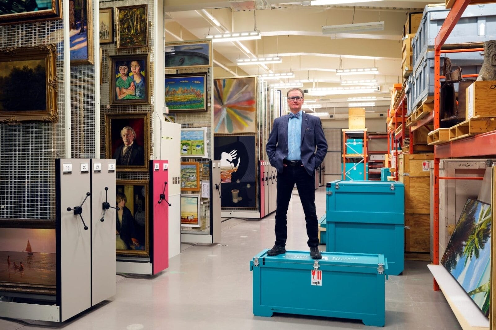 A person wearing a blazer and glasses stands confidently on a teal storage trunk in an organized storage area filled with paintings and other artworks. Whispers from the Vault, the space features neatly arranged shelving and racks filled with various framed art pieces and storage boxes.