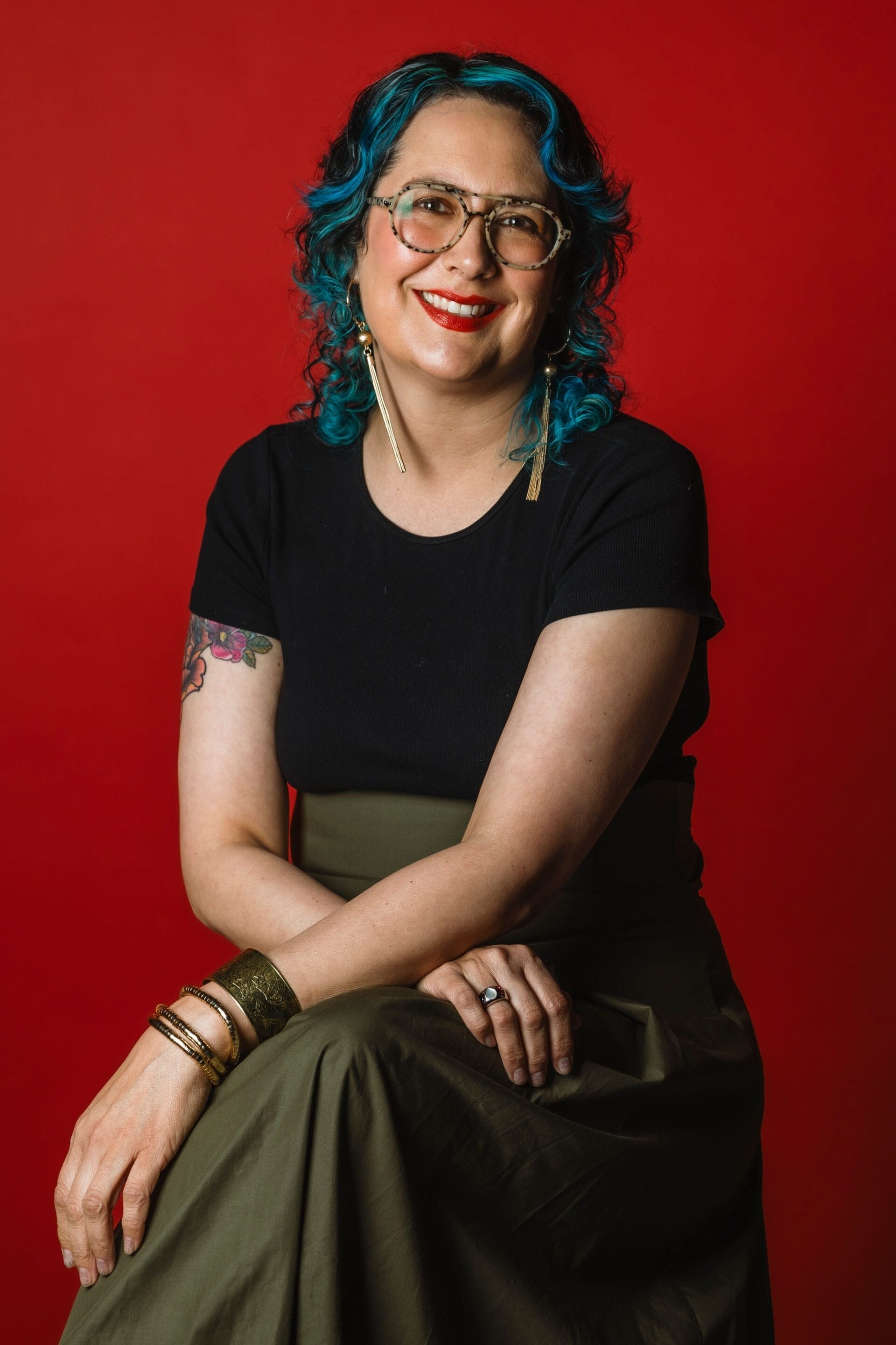 A person with curly blue hair, glasses, and bright red lipstick smiles while sitting against a red background. They wear a black top, olive skirt, hoop earrings, bracelets, and have a floral tattoo on their upper arm.