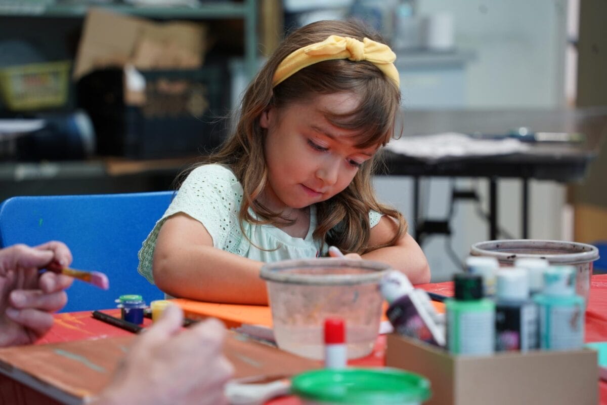 Une jeune fille coiffée d'un bandeau jaune est assise à une table, absorbée par un projet créatif. Du matériel artistique, de la peinture et des pinceaux sont disposés devant elle, tandis qu'un adulte travaille à proximité. Des étagères sont visibles en arrière-plan.