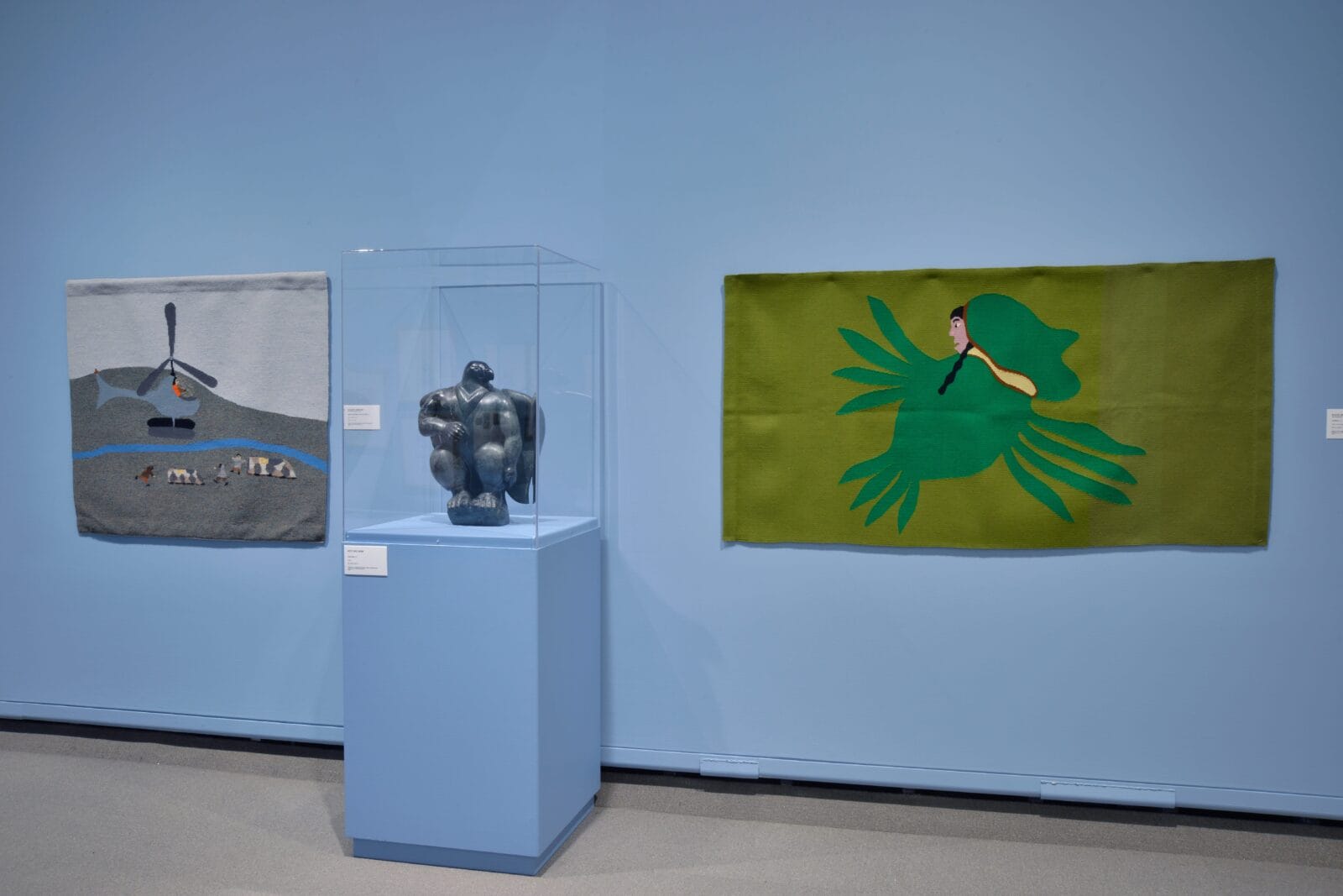 A museum display uses visual storytelling with a dark stone sculpture in a glass case, flanked by vibrant textile artworks on a light blue wall—one depicting a landscape, the other featuring a green bird with a human face.