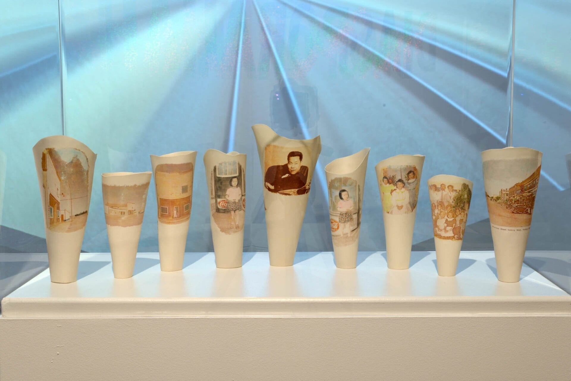 Une rangée de vases en céramique blanche en forme de cône est exposée sur un rebord sur un arrière-plan flou aux lignes convergentes. Chaque vase présente une photographie sépia représentant diverses scènes et personnes, y compris des portraits et des photos de groupe.