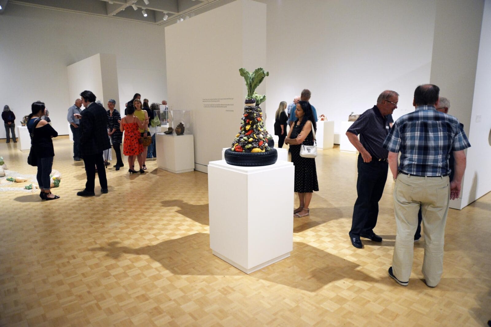 People are observing various art pieces in a spacious art gallery with white walls and wooden floors. A central sculpture on a pedestal made of assorted colorful objects attracts attention. Visitors engage in conversation and appreciate the diverse exhibits.