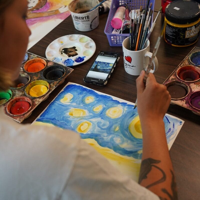 A person creates a Pop Culture Tribute by painting a version of Van Gogh’s “Starry Night” with watercolors, art supplies scattered around, and a phone displaying the artwork reference on the table.