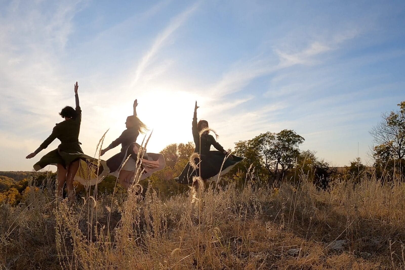 Four people in dresses leap into the air with arms raised in a grassy field at sunset, with tall dry grass and trees in the background, silhouetted against a bright sky.