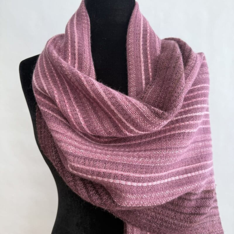 A mauve knit scarf with light pink and white stripes is draped around the neck and shoulders of a black dress form mannequin, set against a plain white background—perfect for gifting or styling during Holiday Bazaart 2025.