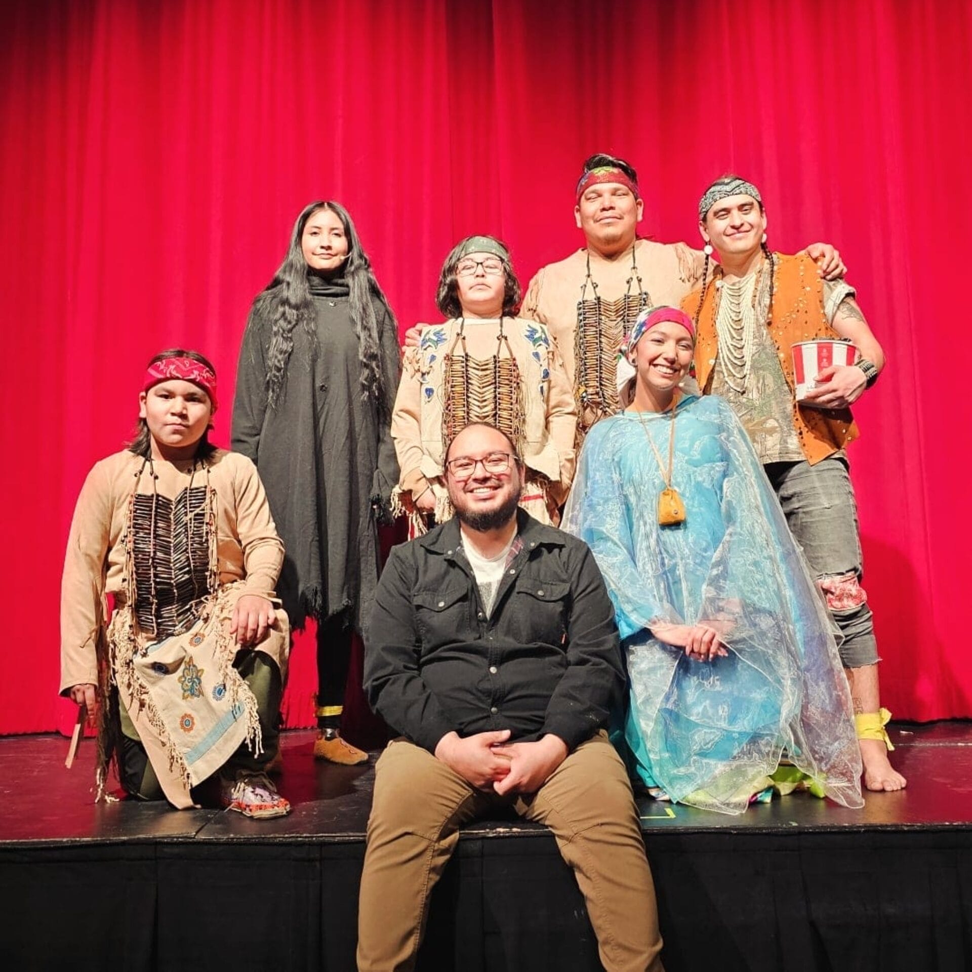 A group of seven people, some in traditional Indigenous attire, pose on a stage in front of red curtains during RAWLCO Free Day. Six stand or kneel, while one sits cross-legged in front, smiling at the camera.