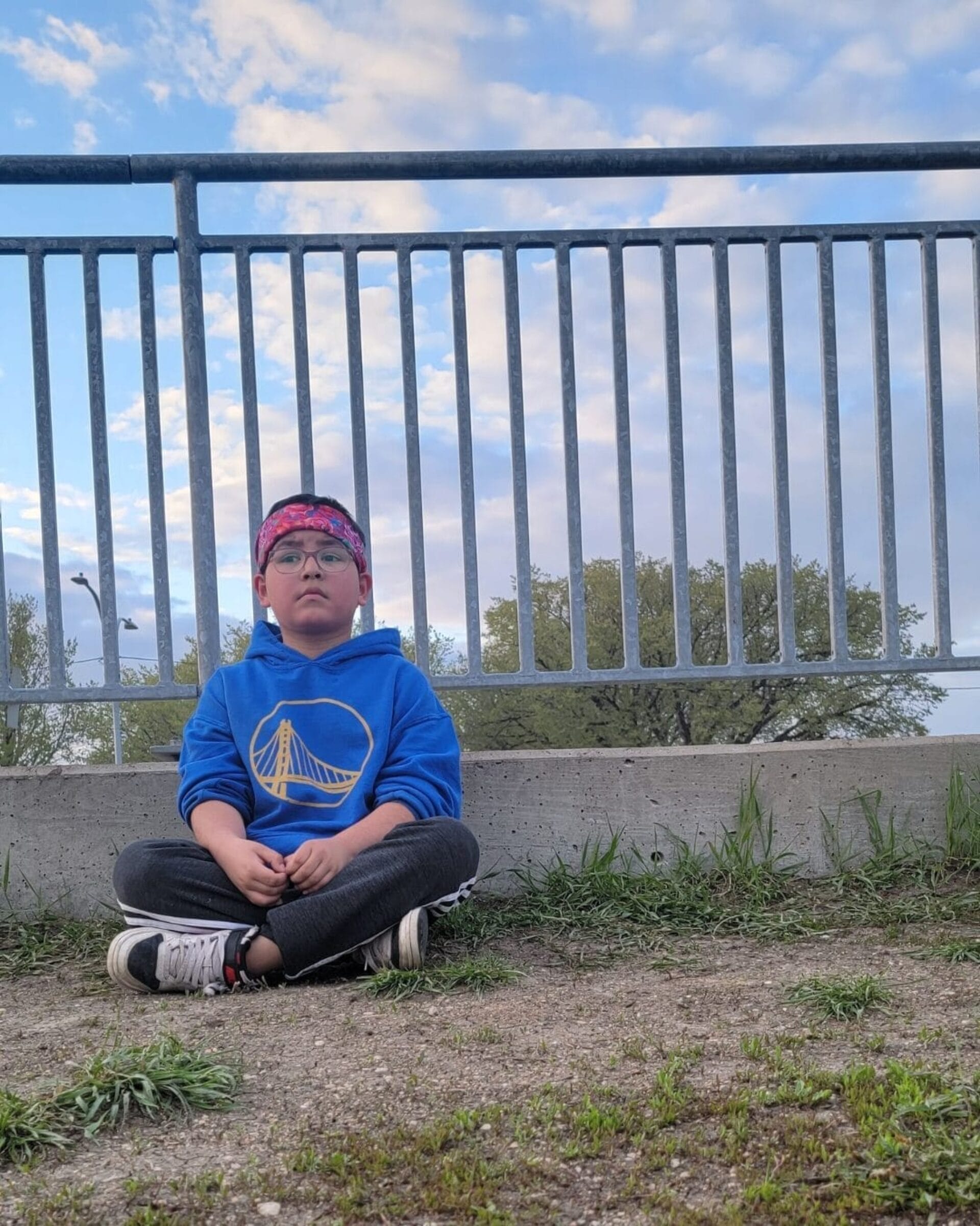 A child wearing glasses, a blue hoodie with a yellow bridge logo, and a pink headband sits cross-legged on the ground in front of a metal fence during RAWLCO Free Day, with trees and a partly cloudy sky in the background.