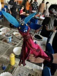A person holds a colorful handmade puppet with blue ears, a blue face, button eyes, and a pink scarf. In the background, children and adults work on arts and crafts at tables covered with newspapers.