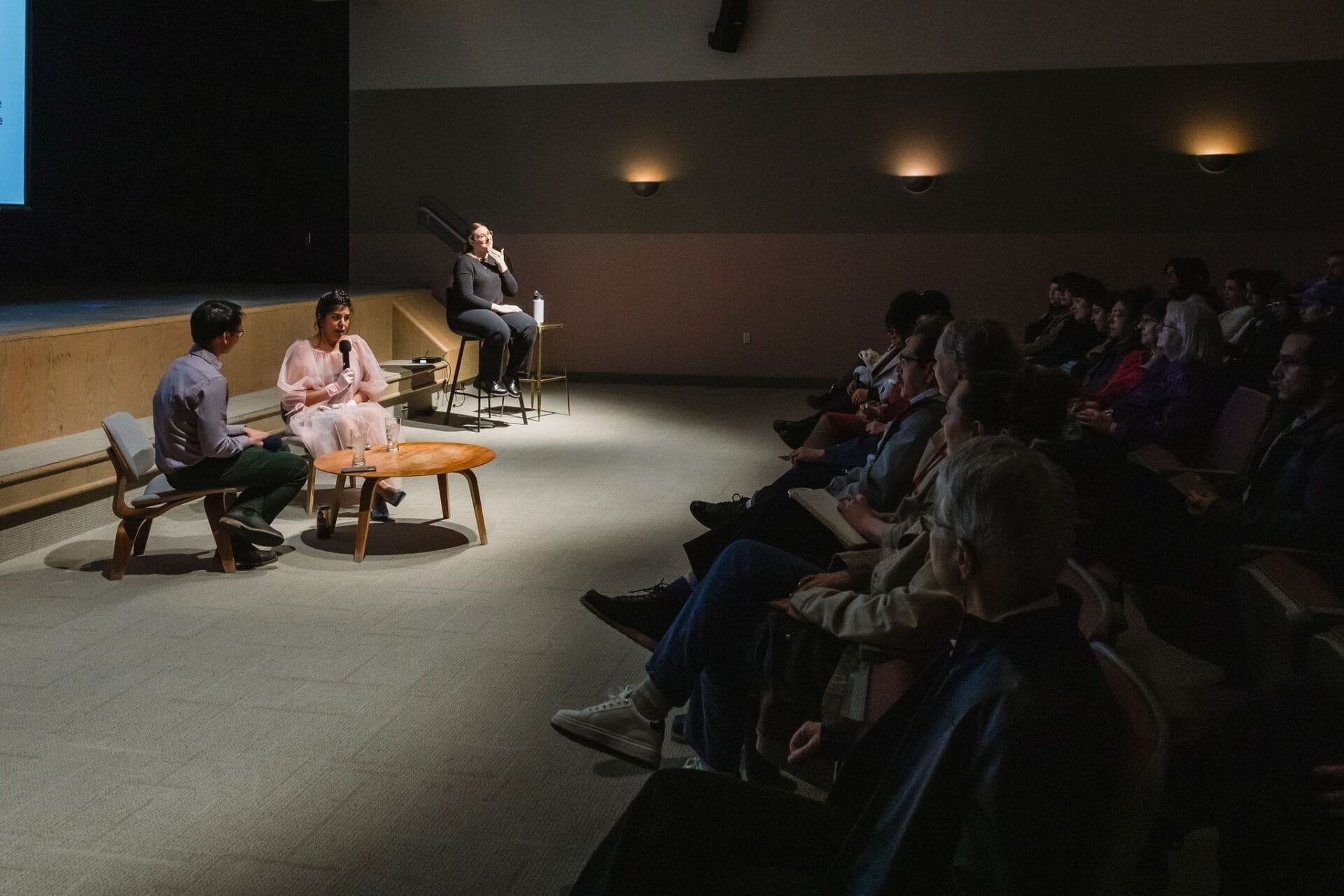Three people participate in a panel discussion on a dimly lit stage facing an audience. Two sit on chairs near a small round table while the third sits on a stool, speaking into a microphone. The audience watches attentively.