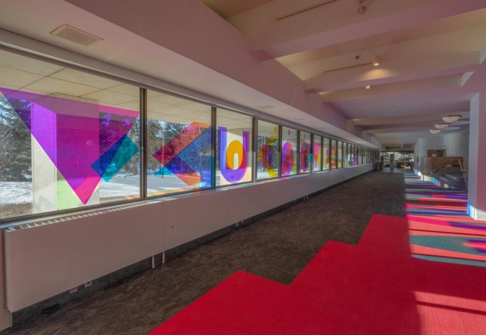 A brightly lit corridor with a long row of windows on the left side, featuring vibrant, geometric stained glass art in various shapes and colors. The sunlight casts colorful patterns onto the floor, which is covered in a mix of red and gray carpeting.