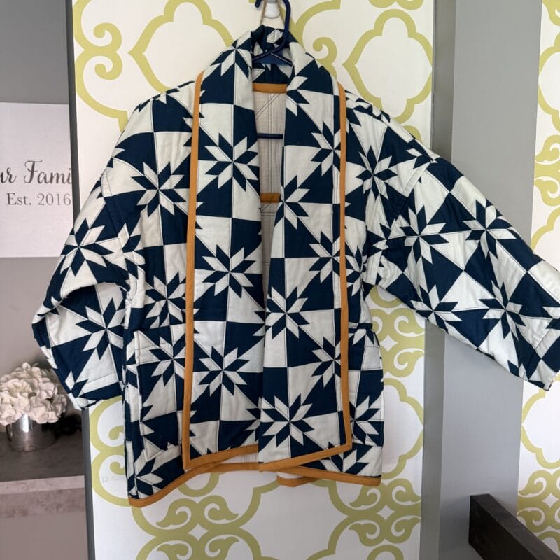 A blue and white patterned jacket with star-like designs and yellow trim hangs on a wall hook against decorative wallpaper, capturing the festive spirit of Holiday Bazaart 2025.