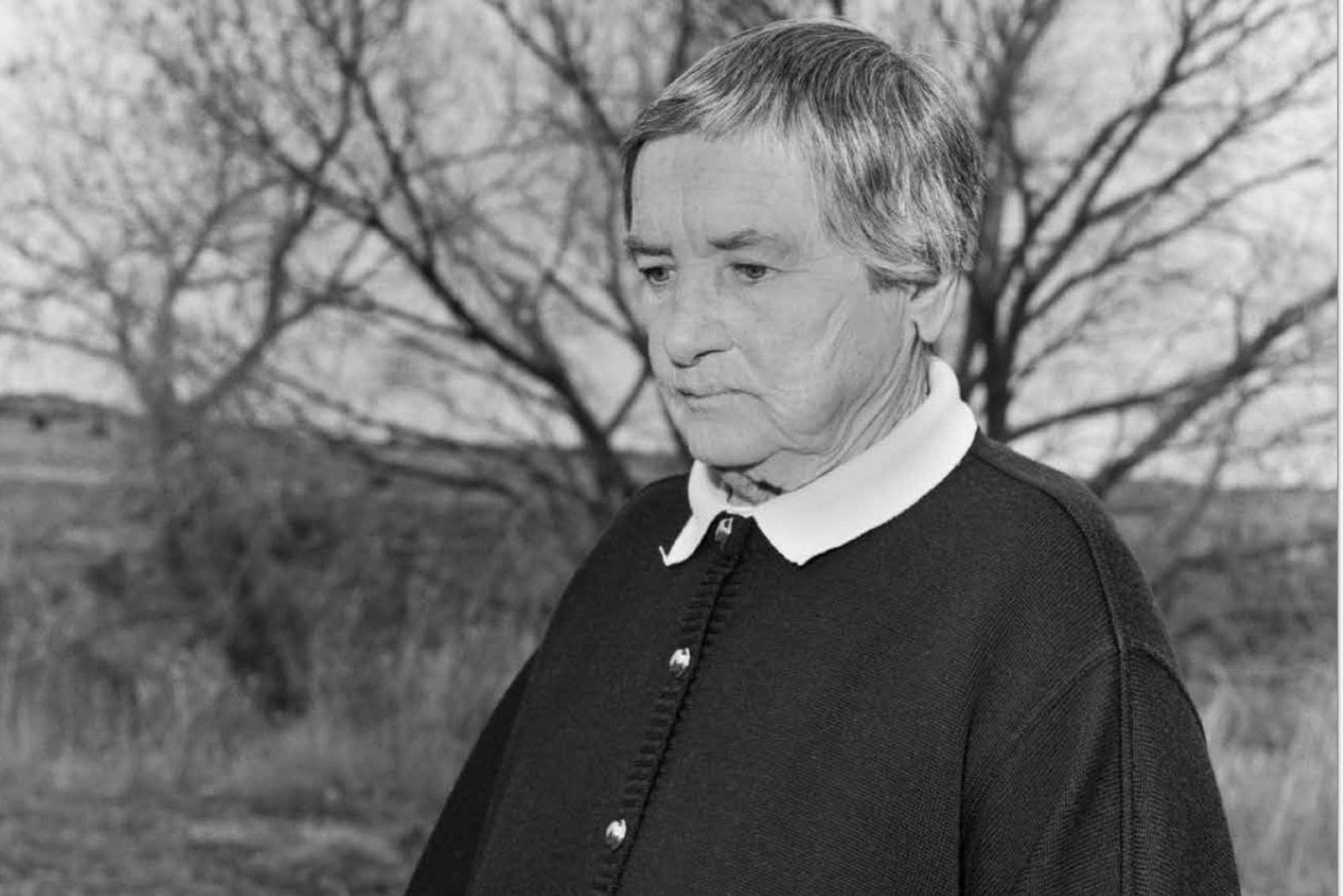 Older man standing facing off to the left with an expressionless face.