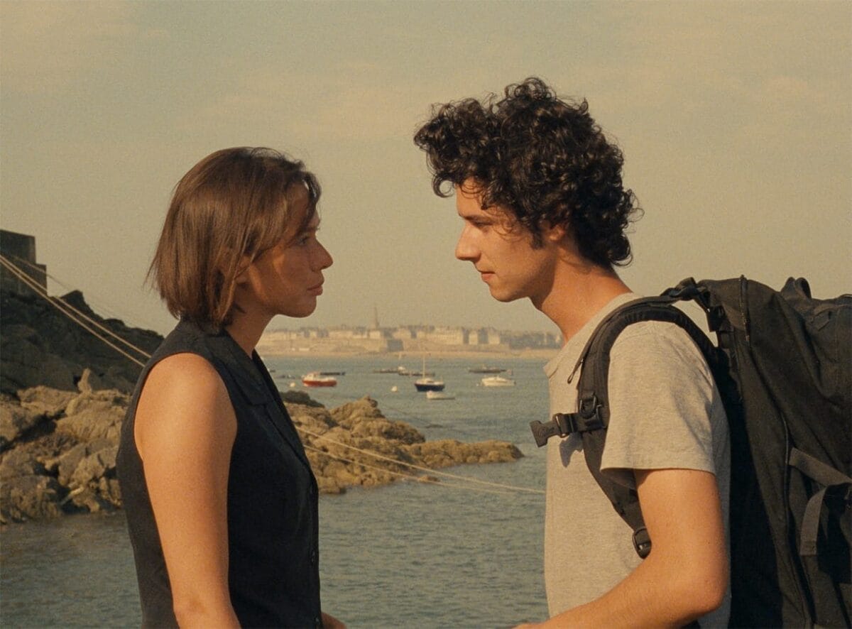 A young woman and man stand facing each other near rocky water’s edge, with boats floating in the background. Bathed in sunlight, this calm scene evokes A Tale of Springtime, as new connections bloom by the gentle shore.