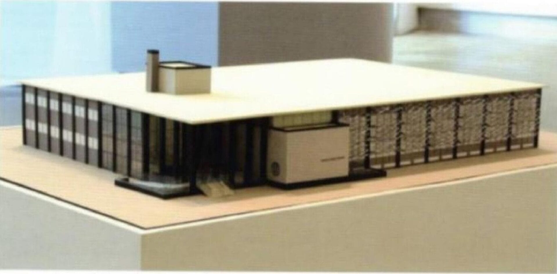 A model of a modernist building with a rectangular structure features a flat roof and dark-tinted glass panels along one side and lattice-like patterns on another. A central glass entrance is visible, accompanied by a small, boxy pinch pot portrait sculpture on the roof.