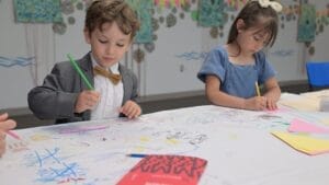 Two young children sit at a table drawing with colored pencils on a large sheet of paper covered in doodles. The boy wears a suit and bow tie, while the girl wears a blue dress. Art supplies are scattered on the table.