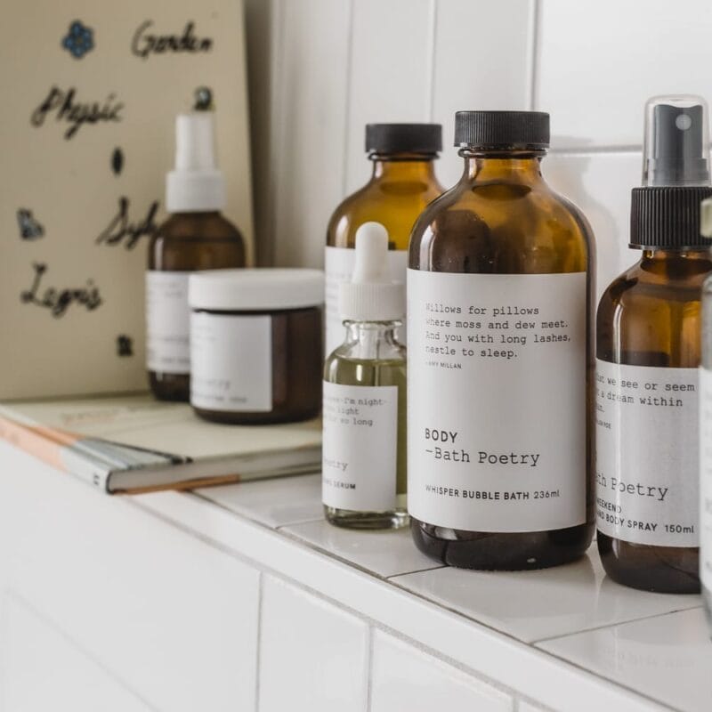 Amidst the serene setting of a bathroom, a collection of brown glass bottles and jars with minimalist white labels sits elegantly on a white shelf. These body and bath essentials, accompanied by a few books, evoke the charm of Holiday Bazaart 2024.