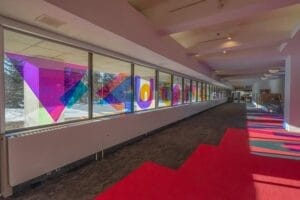 A hallway with large glass windows features colorful geometric shapes, such as triangles and circles, creating vibrant reflections on the floor. The floor has bold red zigzag patterns and the space is filled with natural light.