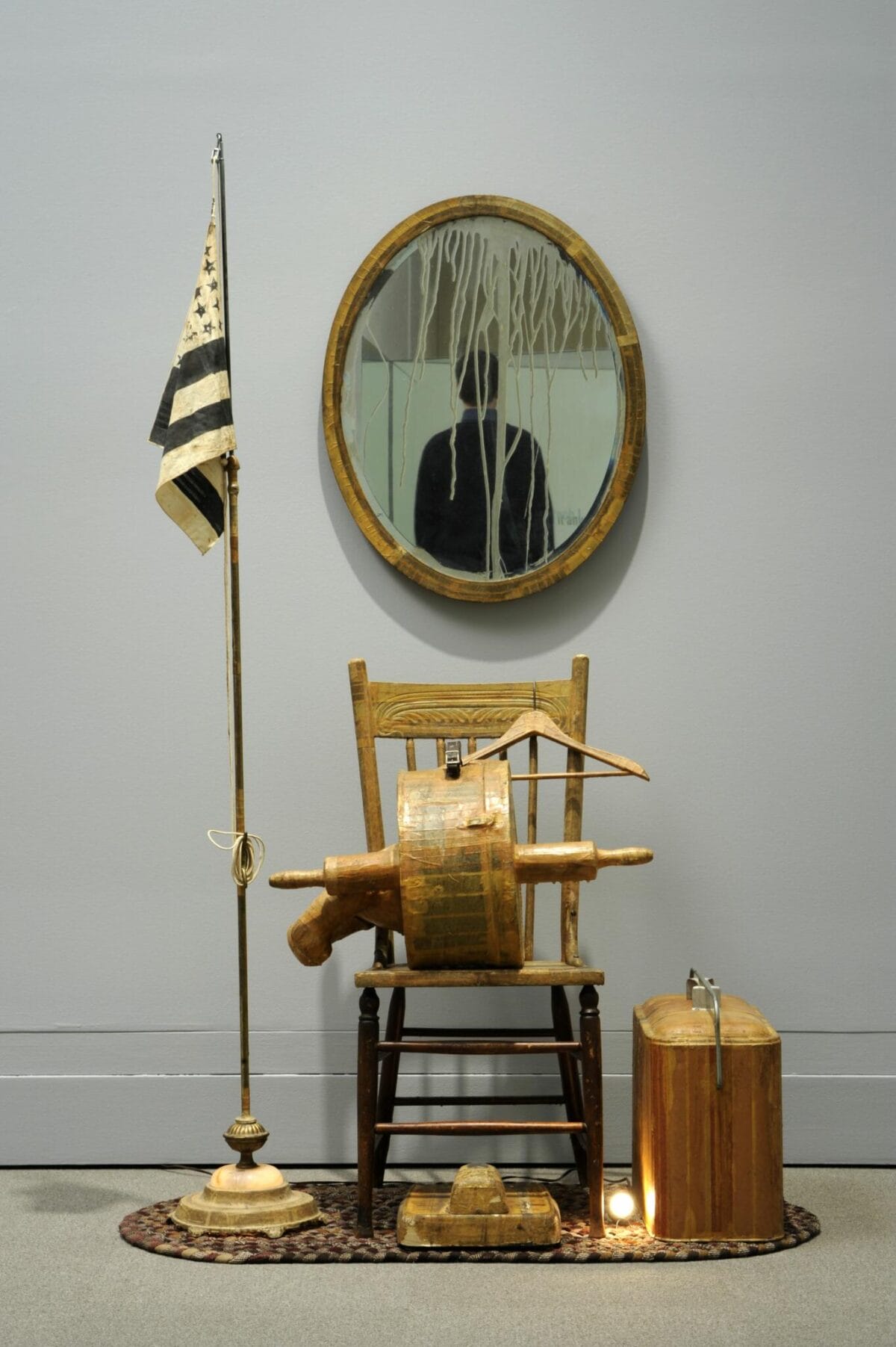 Left: Floor lamp with US flag on top. Center: a straight back wooden chair, coat hanger on the back, a hat box with a rolling pin through it, the back legs of a Teddy Bear stick out of the left side. The hat box is on the chair seat, below the chair is a bathroom scale. Right: a flashing light bulb placed under the chair and a picnic cooler placed on a nylon braided rug on white arborite; behind the chair on the wall is an oval mirror.