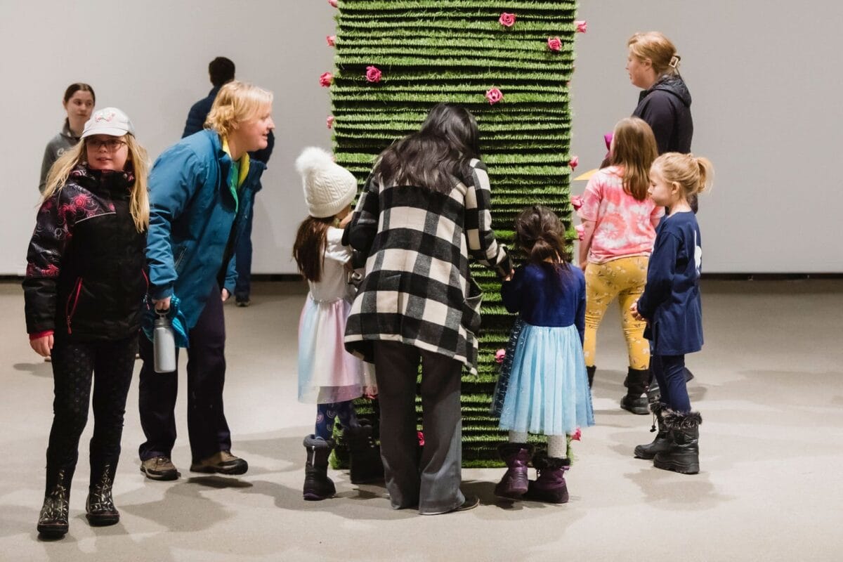 Several children and two adults are gathered around a large, vertical, grass-covered structure with flowers, in a well-lit indoor space. Some children are intently examining the structure, while others stand nearby, seemingly engaged in conversation.