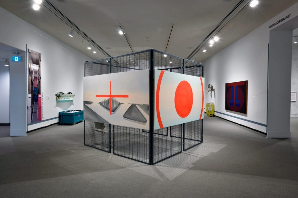 Une installation d'art contemporain dans une galerie présente de grands panneaux avec des motifs abstraits de formes rouges sur un fond. Les pièces sont montées dans un cadre en métal. La salle présente des œuvres d'art supplémentaires et une décoration minimaliste.