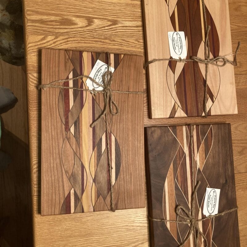 Three wooden cutting boards with intricate patterns of varying wood tones are arranged on a wooden surface. Each board is wrapped in twine, with a tag attached, exuding Holiday Bazaart 2024 charm through their unique swirling designs in contrasting colors.