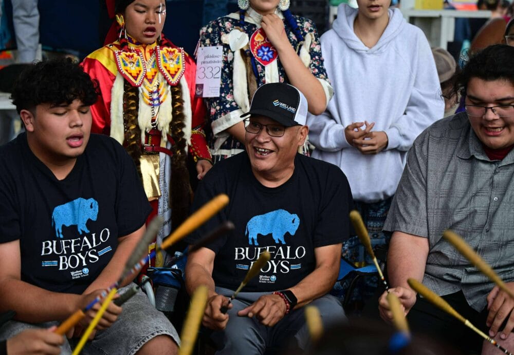 A group of people, including two in “Buffalo Boys” shirts, play drums together at a gathering. Behind them, others wear colorful traditional clothing and regalia, engaging in the event.