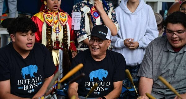 A group of people, including two in “Buffalo Boys” shirts, play drums together at a gathering. Behind them, others wear colorful traditional clothing and regalia, engaging in the event.