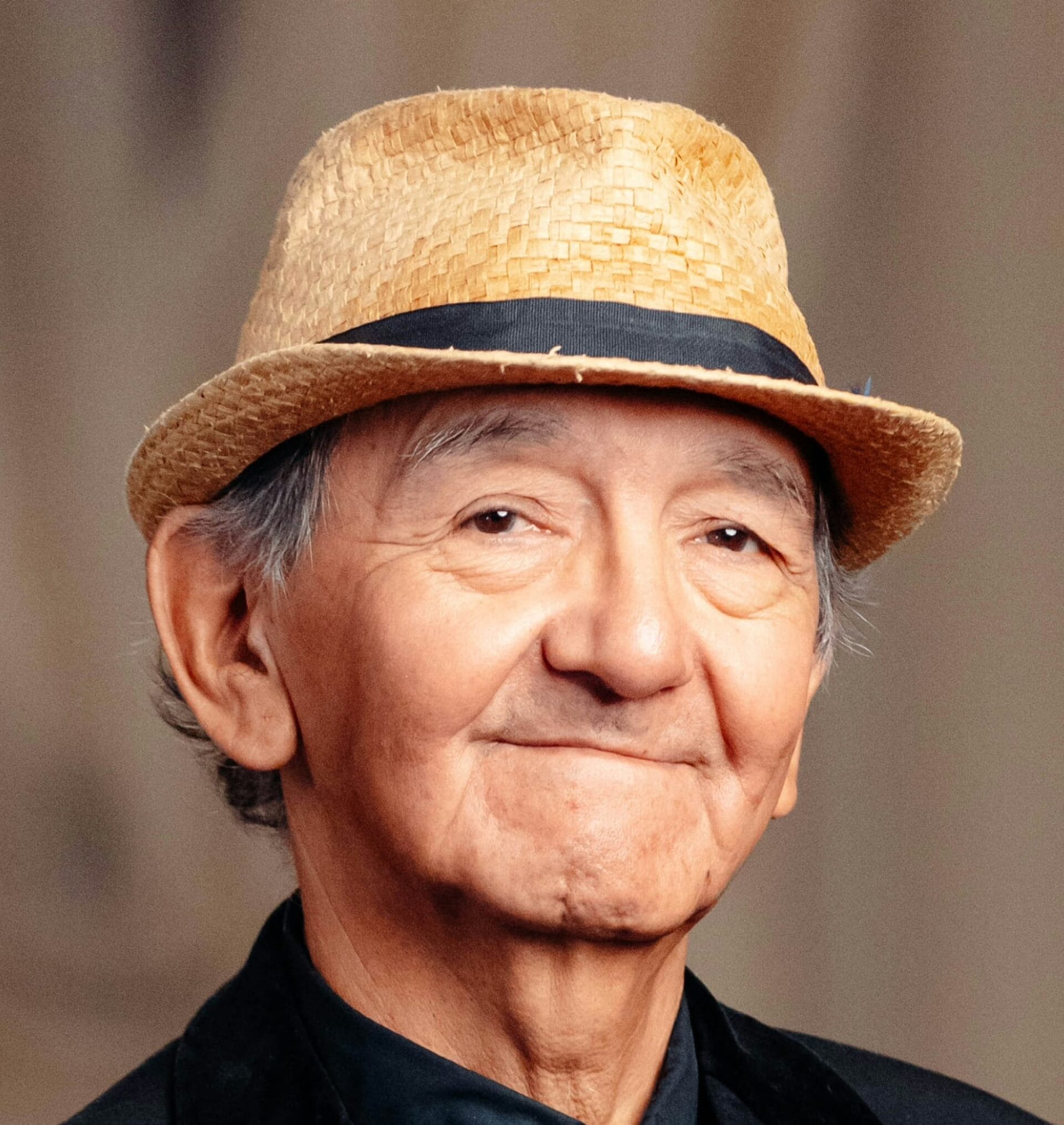 An older man with gray hair wears a straw fedora hat and a dark suit, smiling gently at the camera. The background is blurred and neutral.