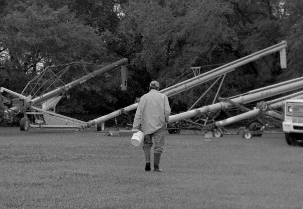 A person wearing a raincoat and boots carries a bucket while walking on grass toward large pieces of farming equipment, with trees and a truck in the background.