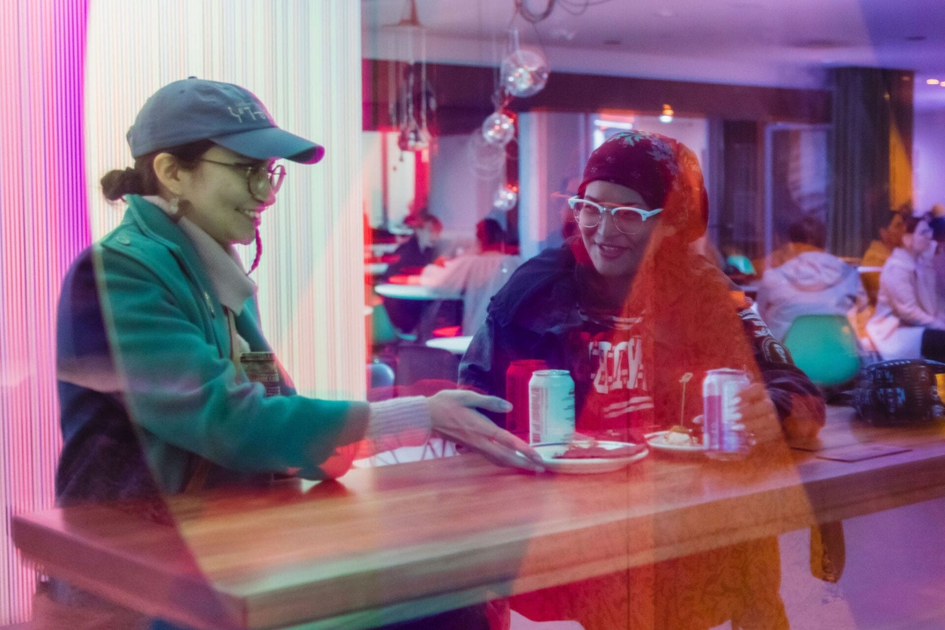 Two people sit at a wooden table indoors, smiling and talking while sharing food and drinks. The scene is viewed through a window, creating colorful reflections and a lively, casual atmosphere. Other people are in the background.