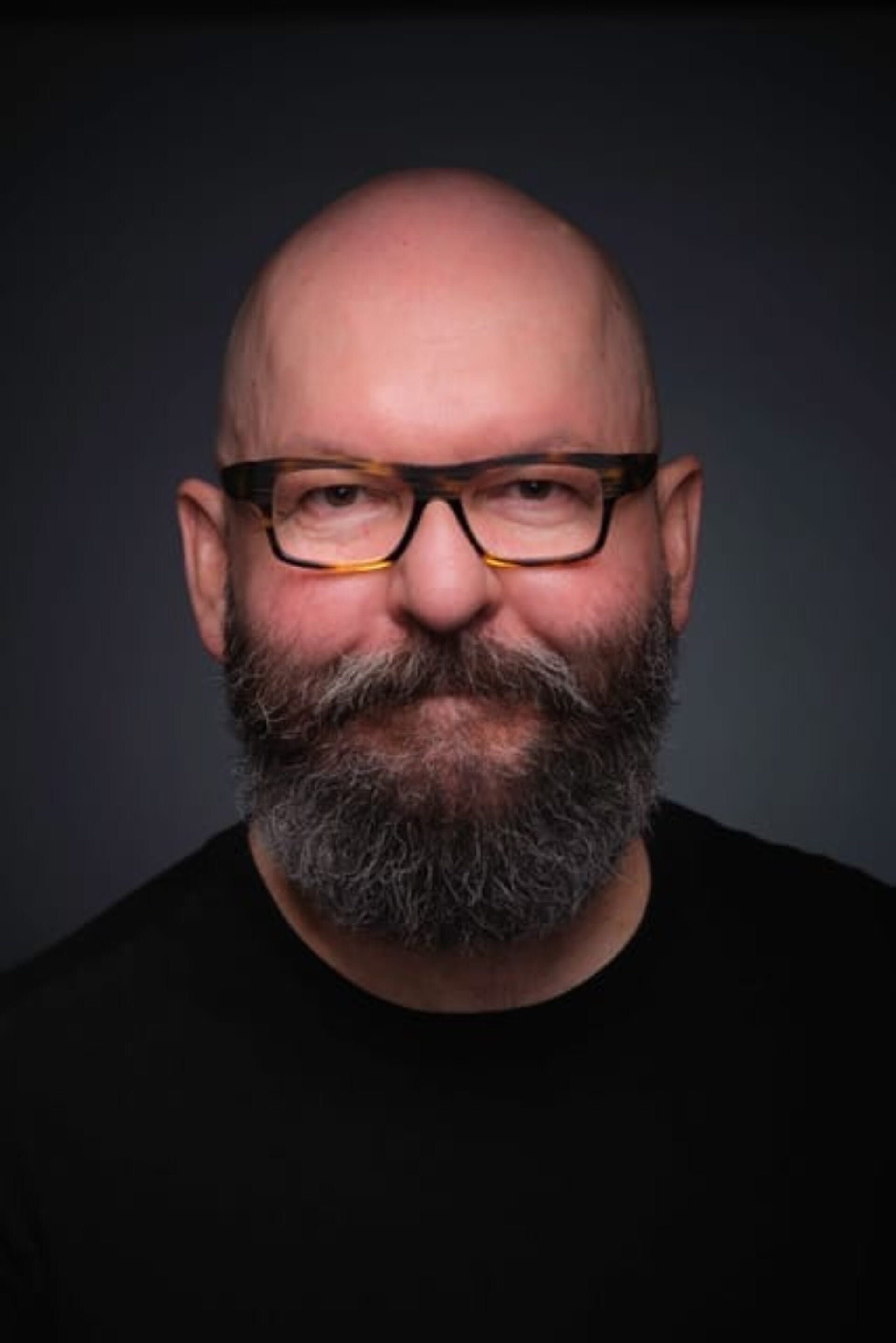 A bald man with a full gray beard and wearing black-framed glasses is smiling slightly. He is dressed in a plain black shirt, and the dark background emphasizes his facial features. He looks like he could be attending a Spring on the Prairie Curator Conversation and Opening Reception event.