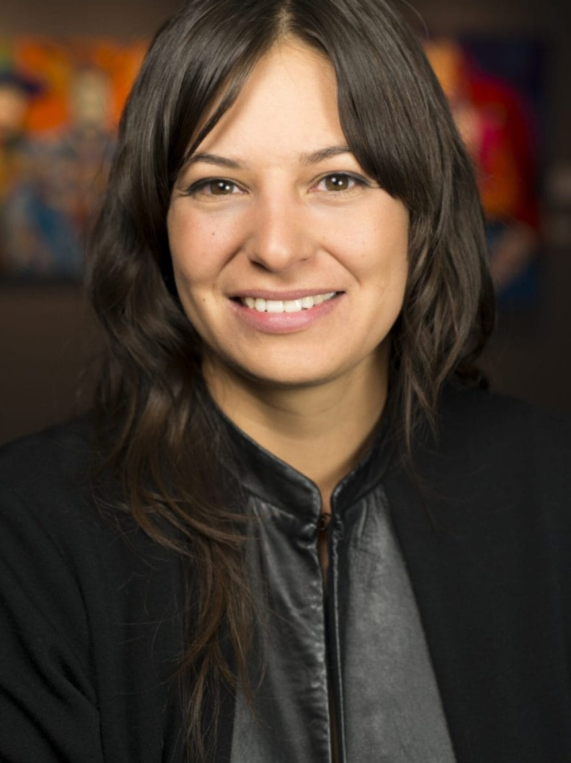 A woman with long brown hair and a friendly smile is wearing a black jacket. The softly blurred background features colorful artwork inspired by Indian Theater.