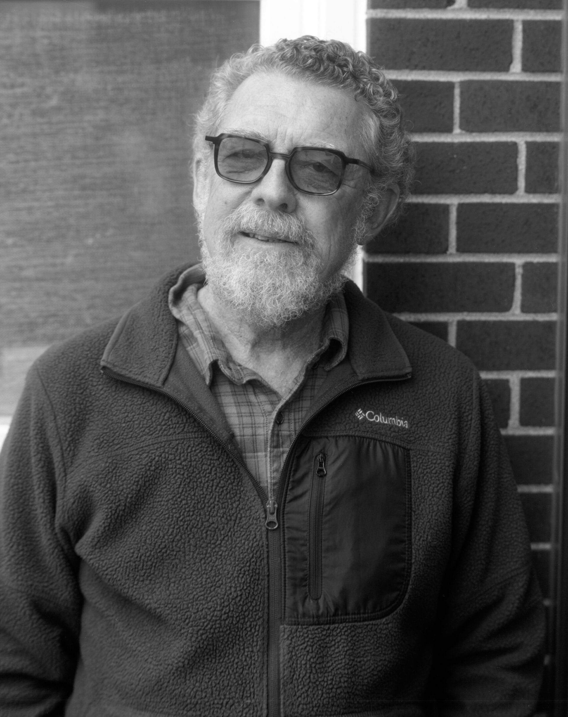 A bearded older man with curly hair, wearing sunglasses, a plaid shirt, and a Columbia fleece jacket, stands in front of a brick wall and window, looking slightly off camera—capturing the contemplative style of Thaddeus Helownia. The photo is black and white.