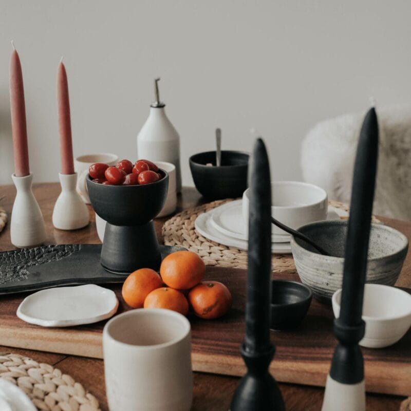 A dining table set with black and white ceramic dishes, tall black and pink candles in holders, a bowl of cherry tomatoes, a few oranges, and woven placemats creates a cozy, minimalist dining scene. Perfectly framed for Holiday gatherings or as inspiration from Bazaart 2024.