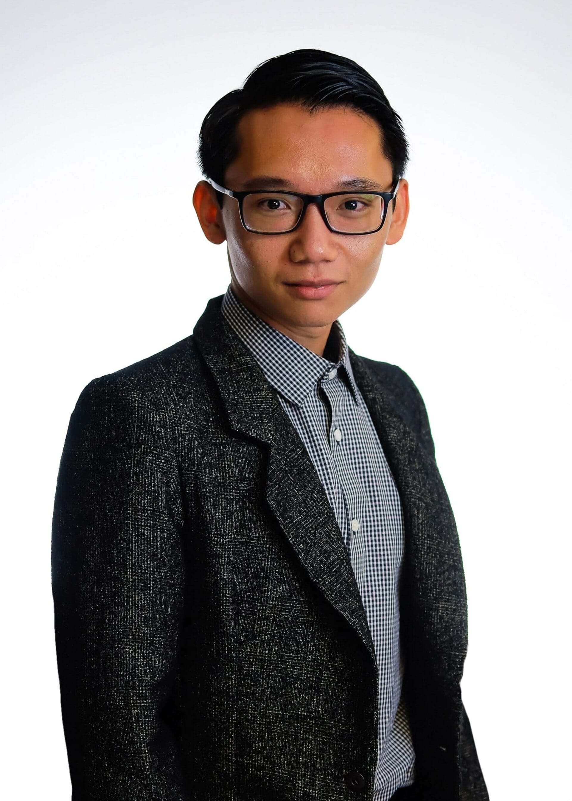 A young person with short, dark hair, wearing glasses, a checkered shirt, and a dark textured blazer stands against a white background, looking confidently at the camera.