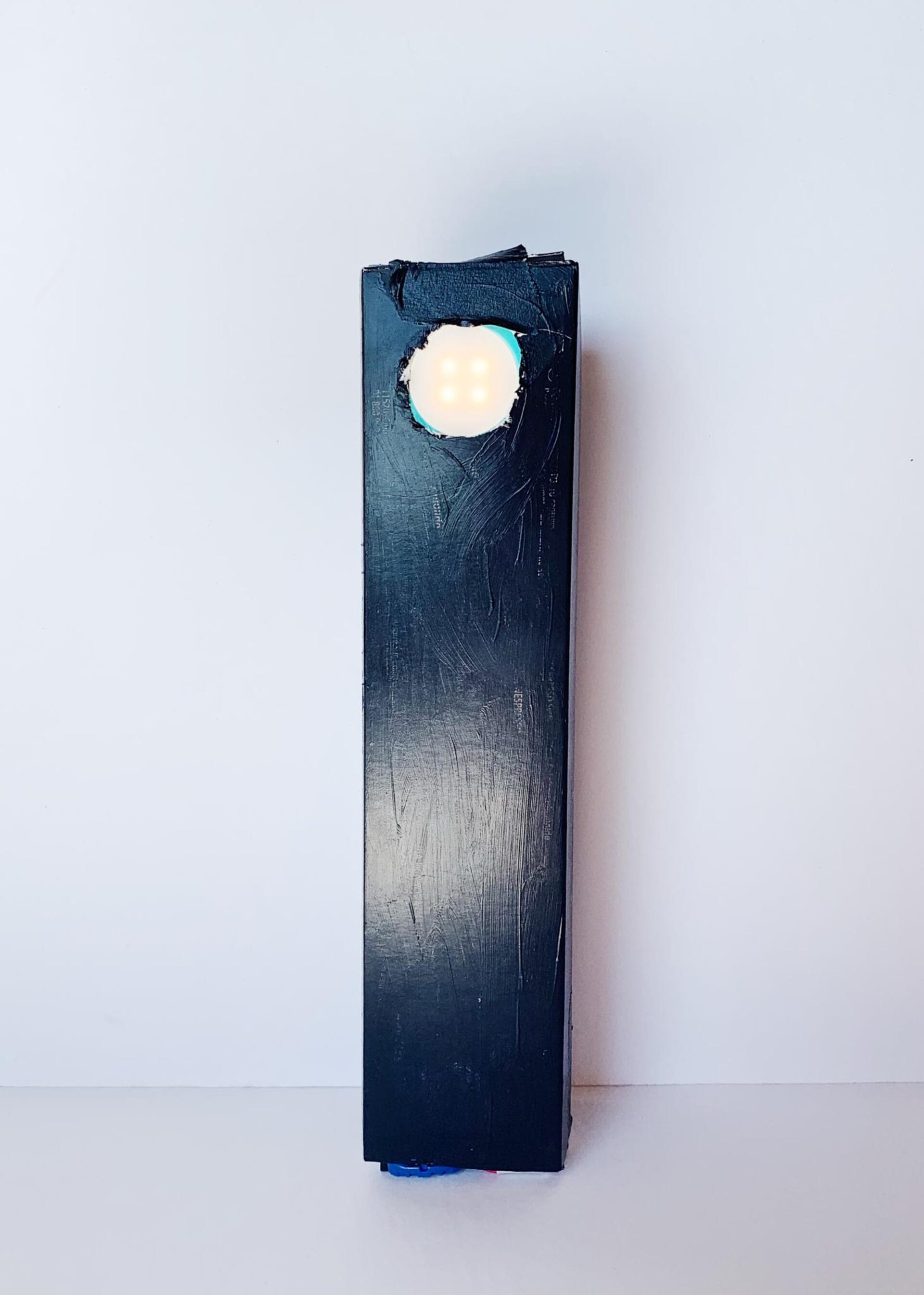 A black rectangular object stands upright against a white background with a rounded light at the top glowing a soft white. The light is central at the top end of the object. The surfaces of the object appear slightly textured or worn.