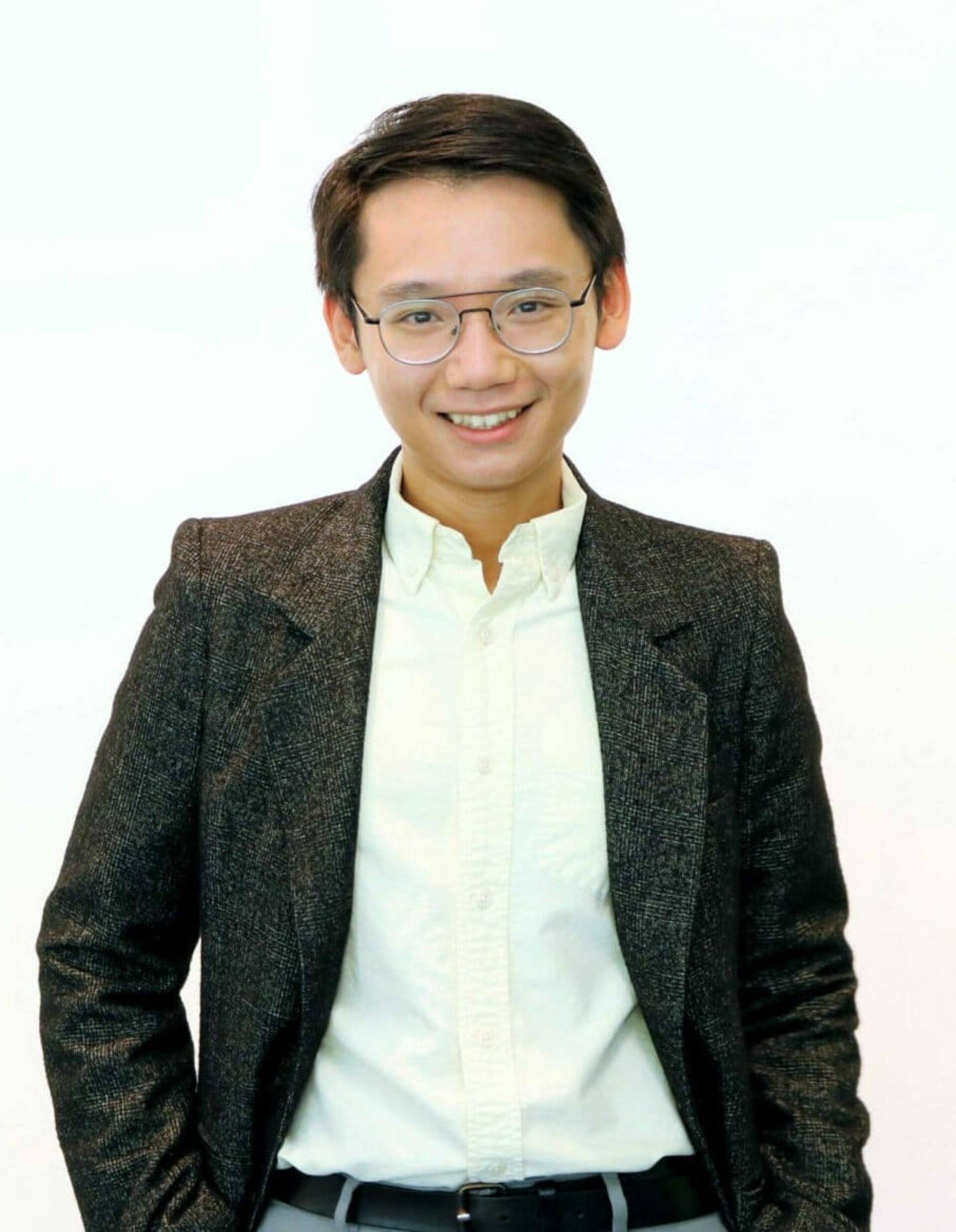 A person with short dark hair, wearing glasses, a dark blazer, and a light-colored button-down shirt smiles at the camera. The background is plain white.