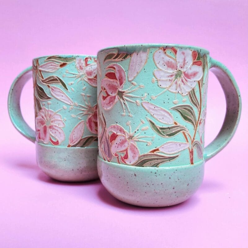 Two ceramic mugs with intricate floral designs in pink and green tones are placed against a pastel pink background. Each mug features detailed flowers and leaves, creating a delicate and artistic appearance, perfect for adding a touch of charm to your Holiday Bazaart 2024 collection.