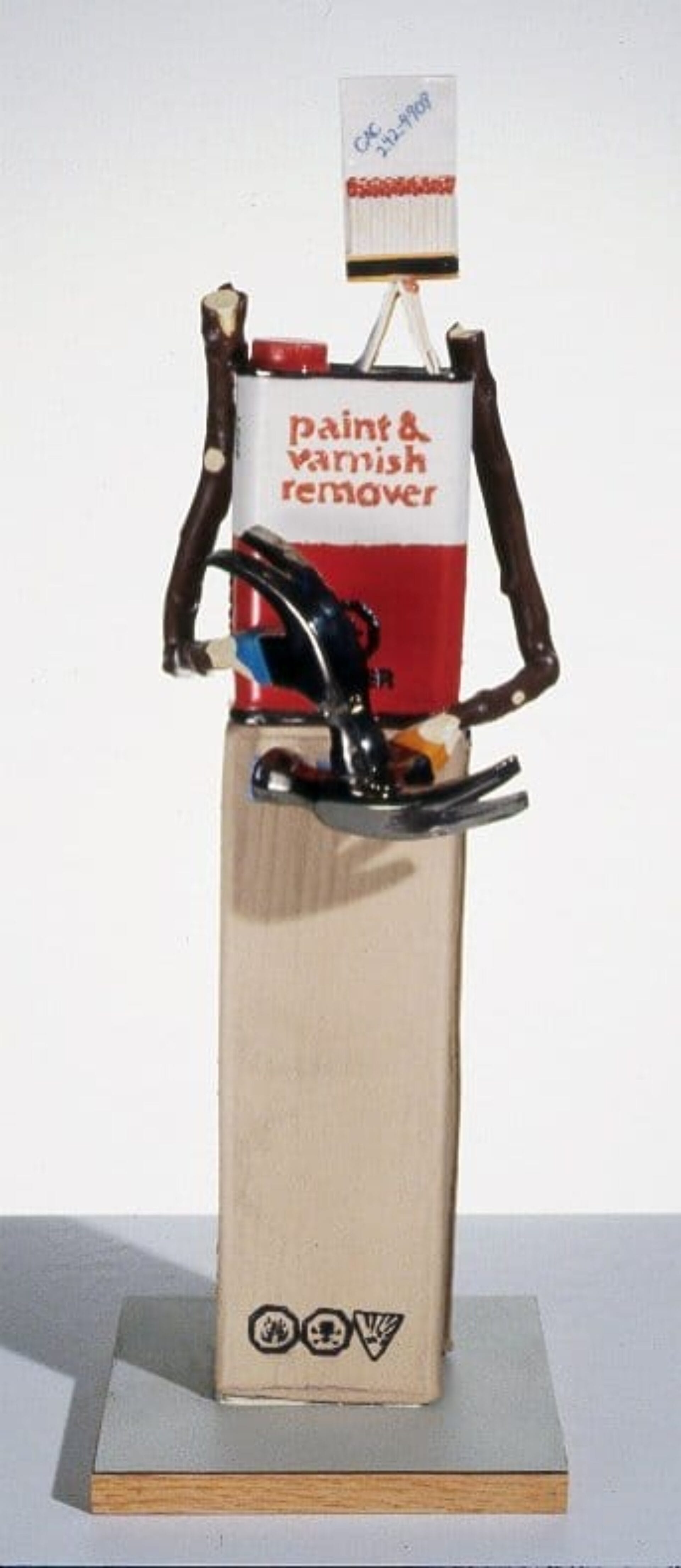 A quirky sculpture depicts a humanoid figure made from a paint and varnish remover can, a cardboard base, wire arms, and metal claw-like hands. The head is a small box with 