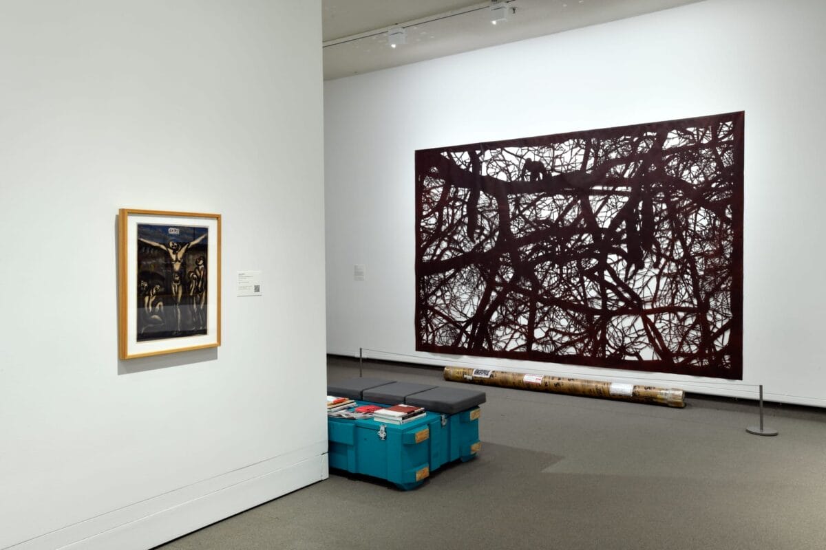Une galerie d'art moderne présentant deux œuvres d'art : un petit tableau encadré sur un mur blanc et une grande œuvre abstraite aux motifs sombres complexes. La salle dispose d'un éclairage doux et d'un banc avec des livres au premier plan.