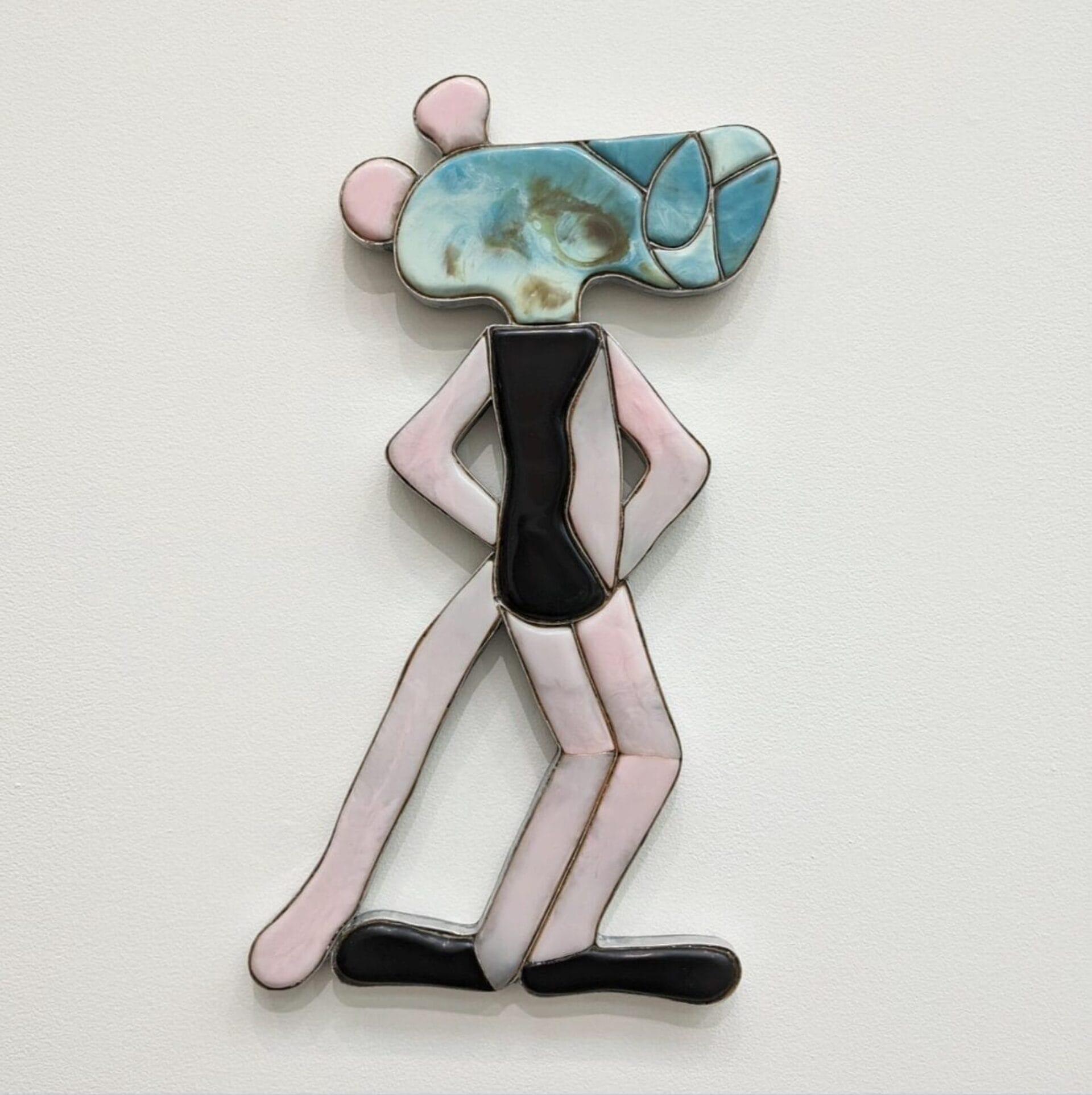 A stylized, abstract figure inspired by pop culture tributes stands against a white background, crafted from pastel-colored and black materials with exaggerated limbs, a narrow torso, and a large blue-tinged head with round ears.