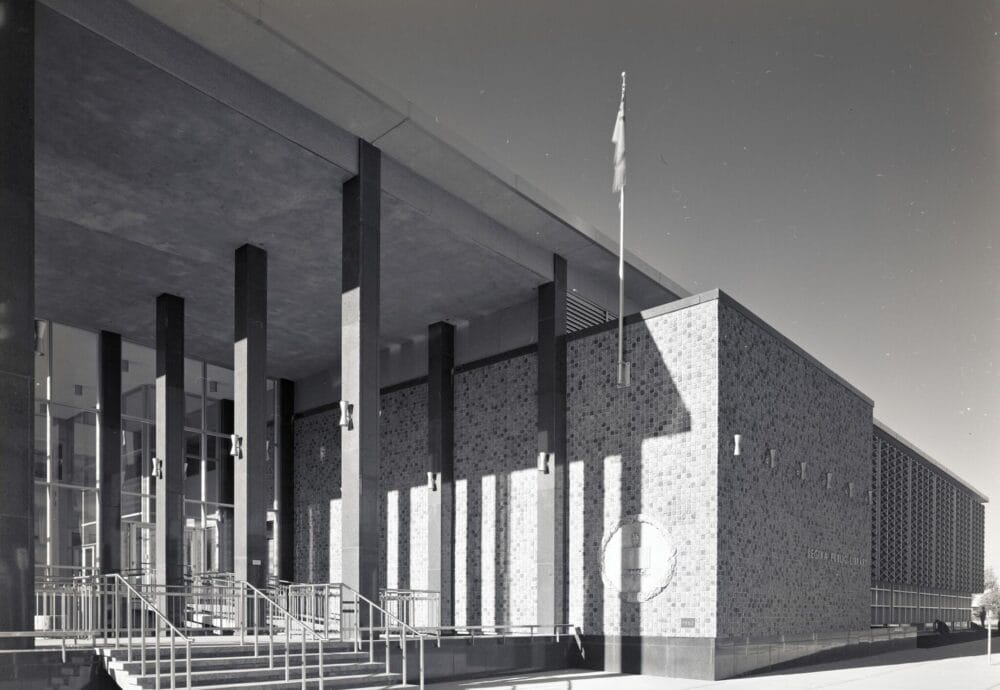 Black and white image of a modernist-style public building with large vertical pillars, a flat roof, and a brick exterior. The structure features a prominent entrance with a glass facade, multiple steps, and handrails. A flagpole is visible near the entrance, adding to the serene atmosphere reminiscent of Spring on the Prairies.