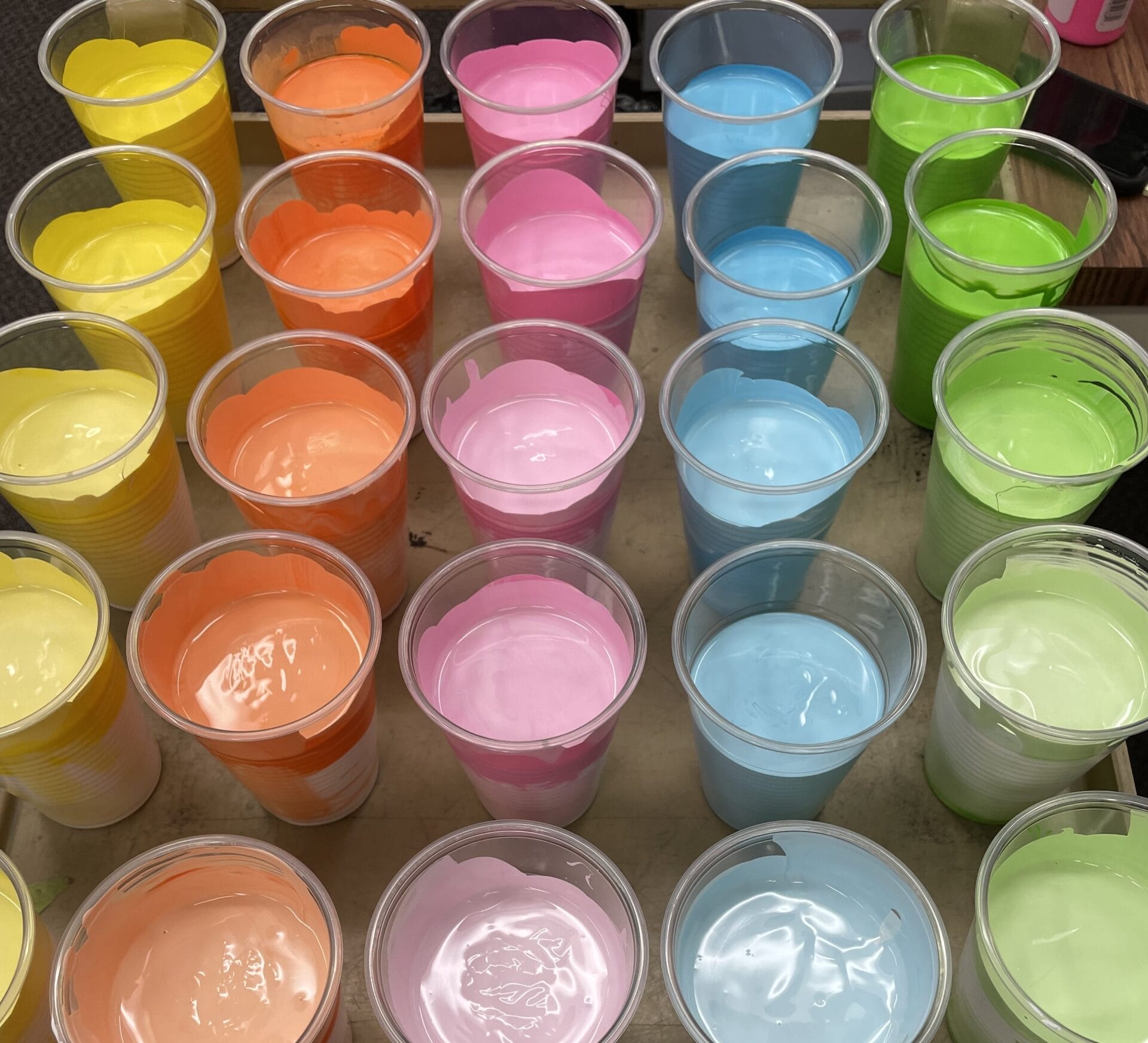 Des gobelets en plastique transparent remplis de peinture colorée dans les tons jaune, orange, rose, bleu et vert sont disposés en rangs bien ordonnés sur une surface plane.