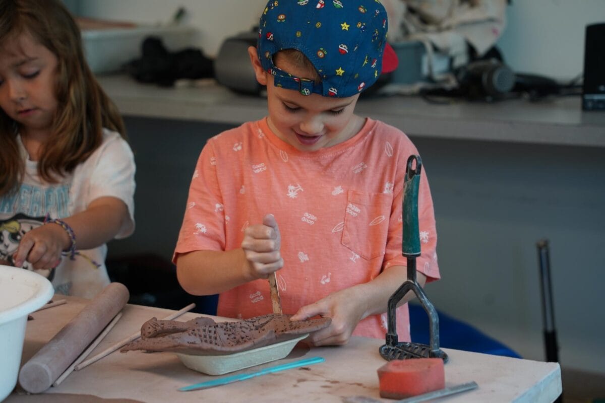 Un jeune enfant portant une casquette bleue et une chemise rose façonne l'argile avec un outil, assis à une table, tandis qu'un autre enfant travaille à proximité. Divers outils et un rouleau à pâtisserie sont également posés sur la table.